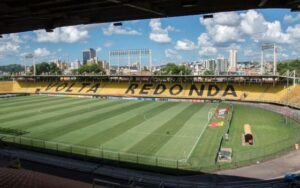 Vista panorâmica do Estádio Raulino de Oliveira em Volta Redonda, com o campo de futebol e as arquibancadas sob um céu azul.